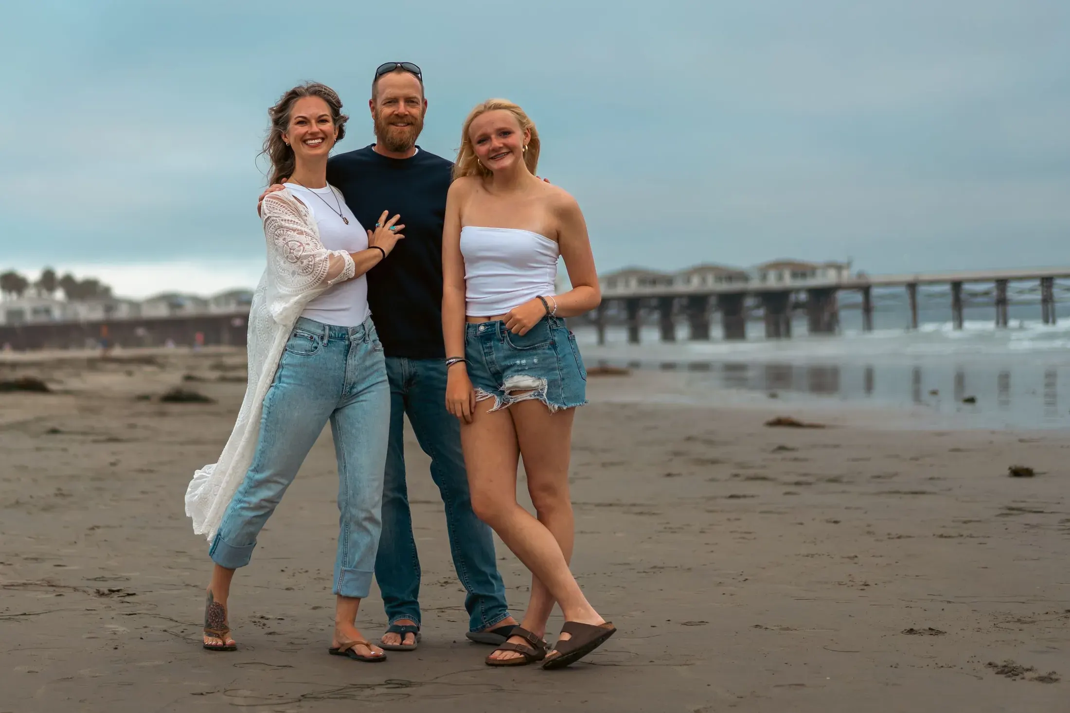Family of three smiling on the beach near a pier