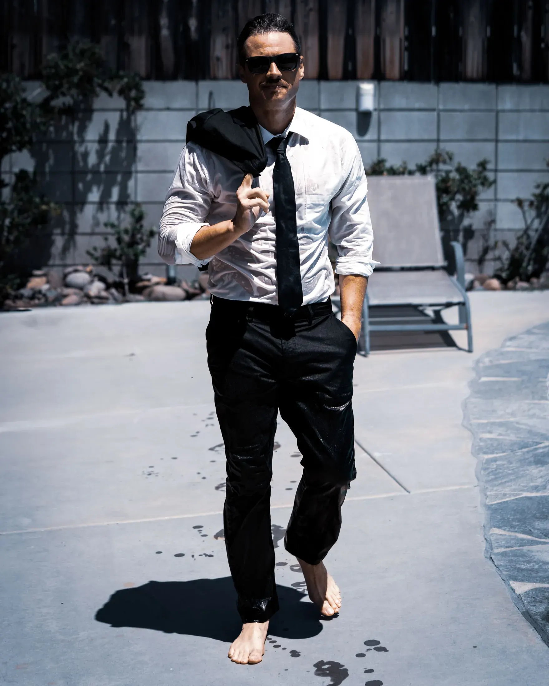 Barefoot man in a white shirt and loose tie walking by a pool